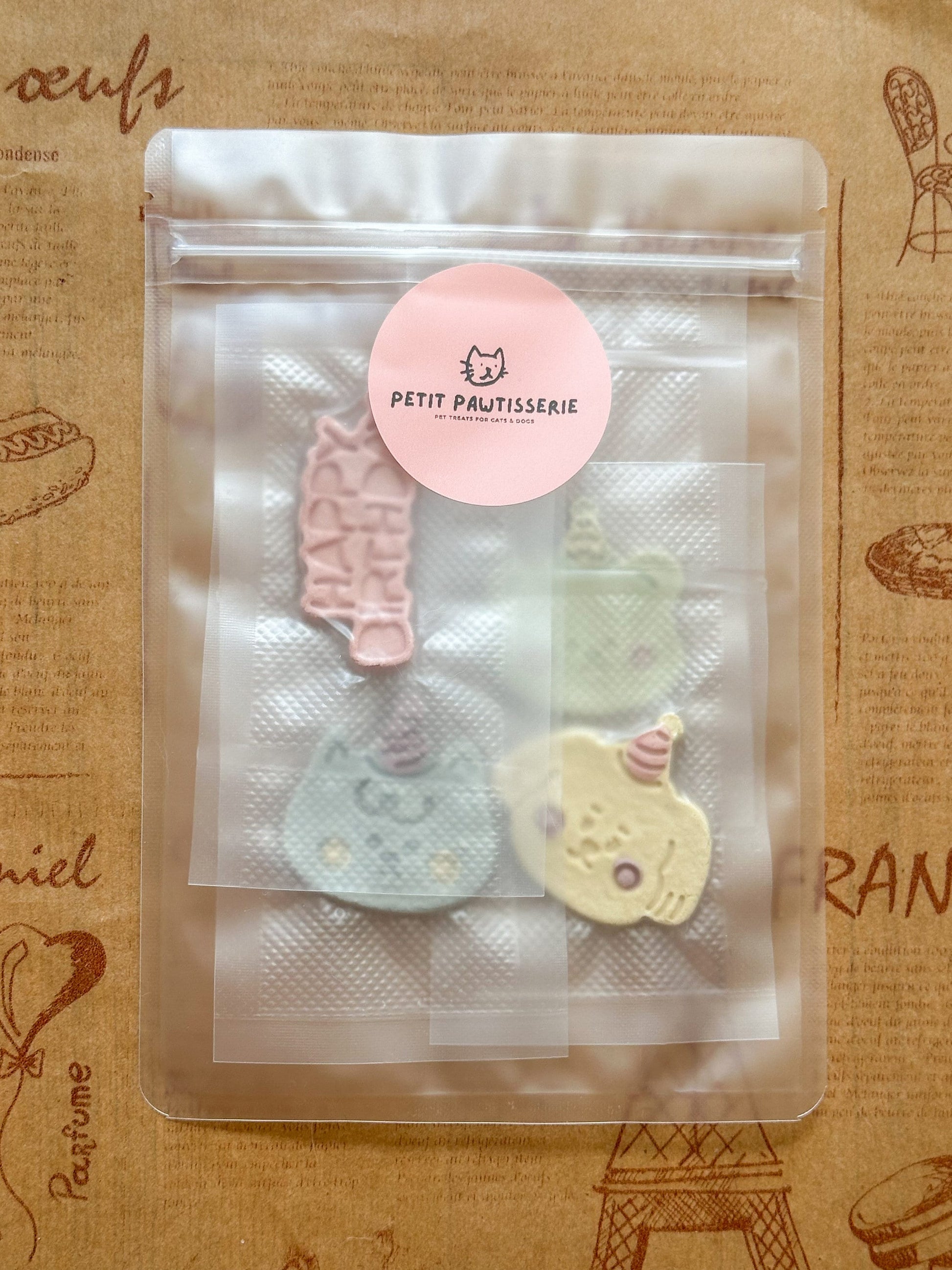 A clear plastic bag of pastel-colored pet treats. The treats are shaped like cats and other animals. A pink circle sticker on the bag says 'PETIT PAWTISSERIE Pet Treats for Cats & Dogs'.
