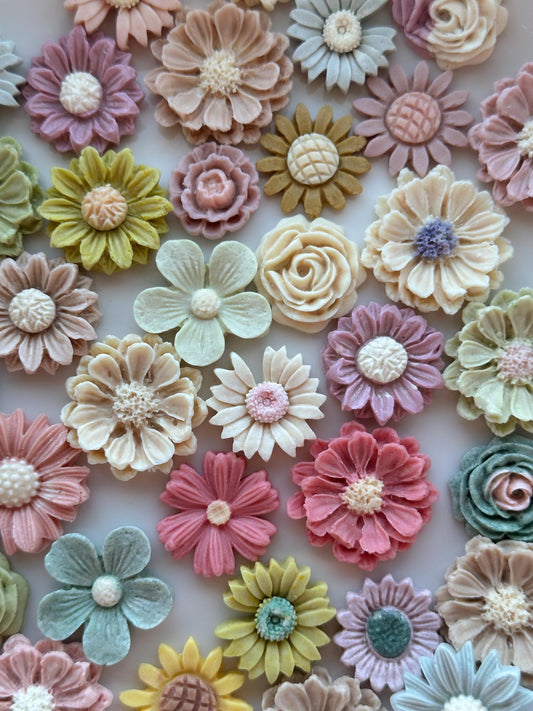 A collection of colorful handmade goat milk flower pet treats in various shapes and sizes. The flowers are in shades of green, pink, purple, and white.