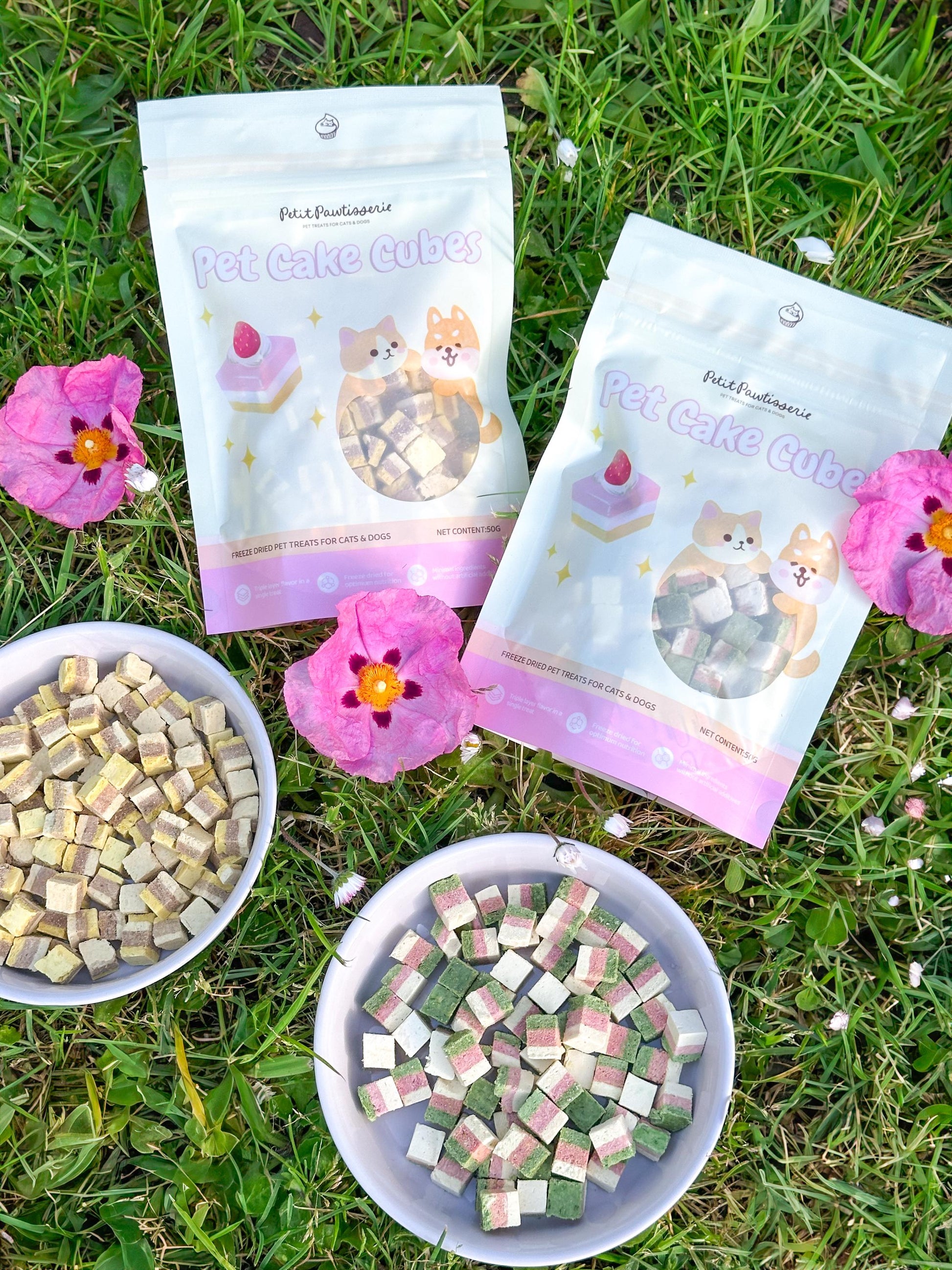 Two bags of pet cake cubes, one labeled 'Pet Cake Cubes' and the other 'Pet Cake Cube.' The bags are on a bed of green grass with pink flowers. The bags are white with pink and yellow accents. The bags feature cartoon images of dogs and cakes. Two bowls of pet cake cubes are in the foreground.  Pawtisserie freeze-dried cake cube pet treats for cats and dogs – soft bite sized snacks with 3 layers of flavor