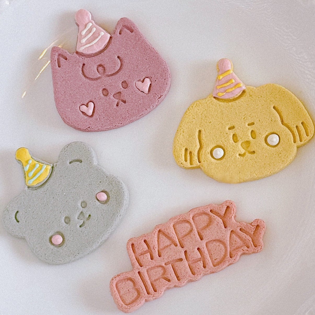 three animal-shaped cookies and a Happy Birthday banner cookie.  The cookies are decorated with icing and feature pastel colors: pink cat, yellow dog, blue bear, and a pink Happy Birthday banner. Each cookie has a small party hat.  The cookies are suitable for pet birthday celebrations and parties.