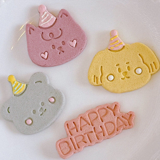 three animal-shaped cookies and a Happy Birthday banner cookie.  The cookies are decorated with icing and feature pastel colors: pink cat, yellow dog, blue bear, and a pink Happy Birthday banner. Each cookie has a small party hat.  The cookies are suitable for pet birthday celebrations and parties.