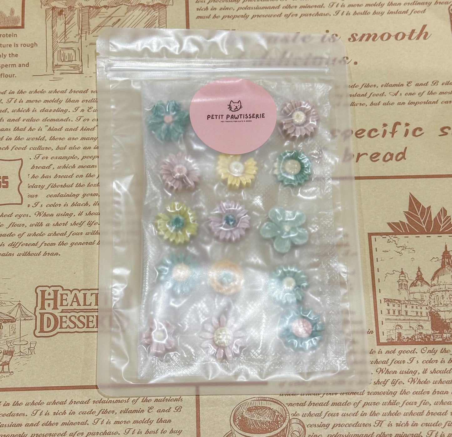 A clear plastic bag with a white background containing handmade goat milk flower pet treats. The flowers are in various shades of green, pink, and yellow. The bag has a small pink label that reads &#39;PETIT PAWTISSERIE&#39;.