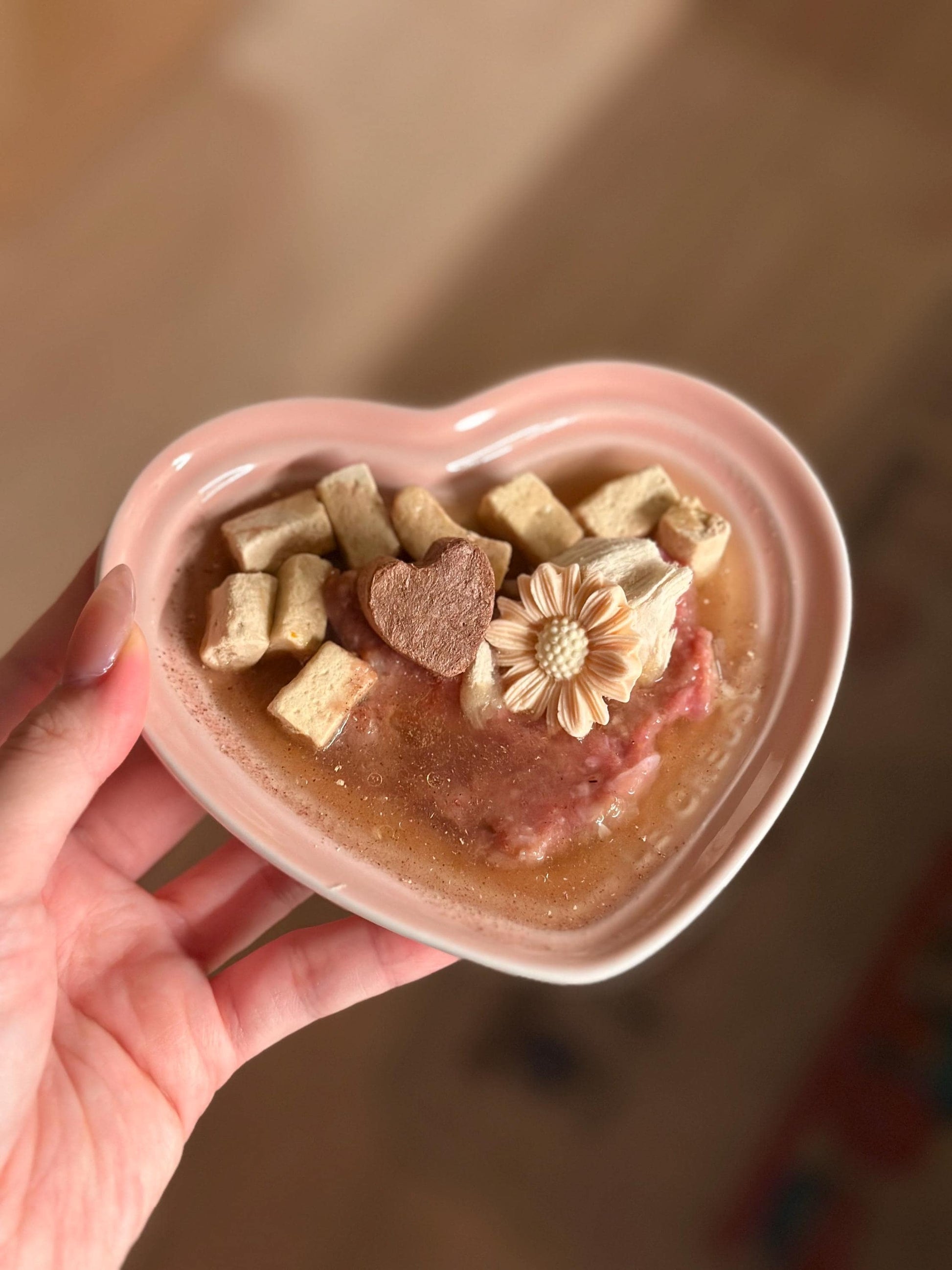 A pink heart-shaped bowl filled with brown and beige pet food, a brown heart-shaped treat, and a pink flower-shaped pet treat.