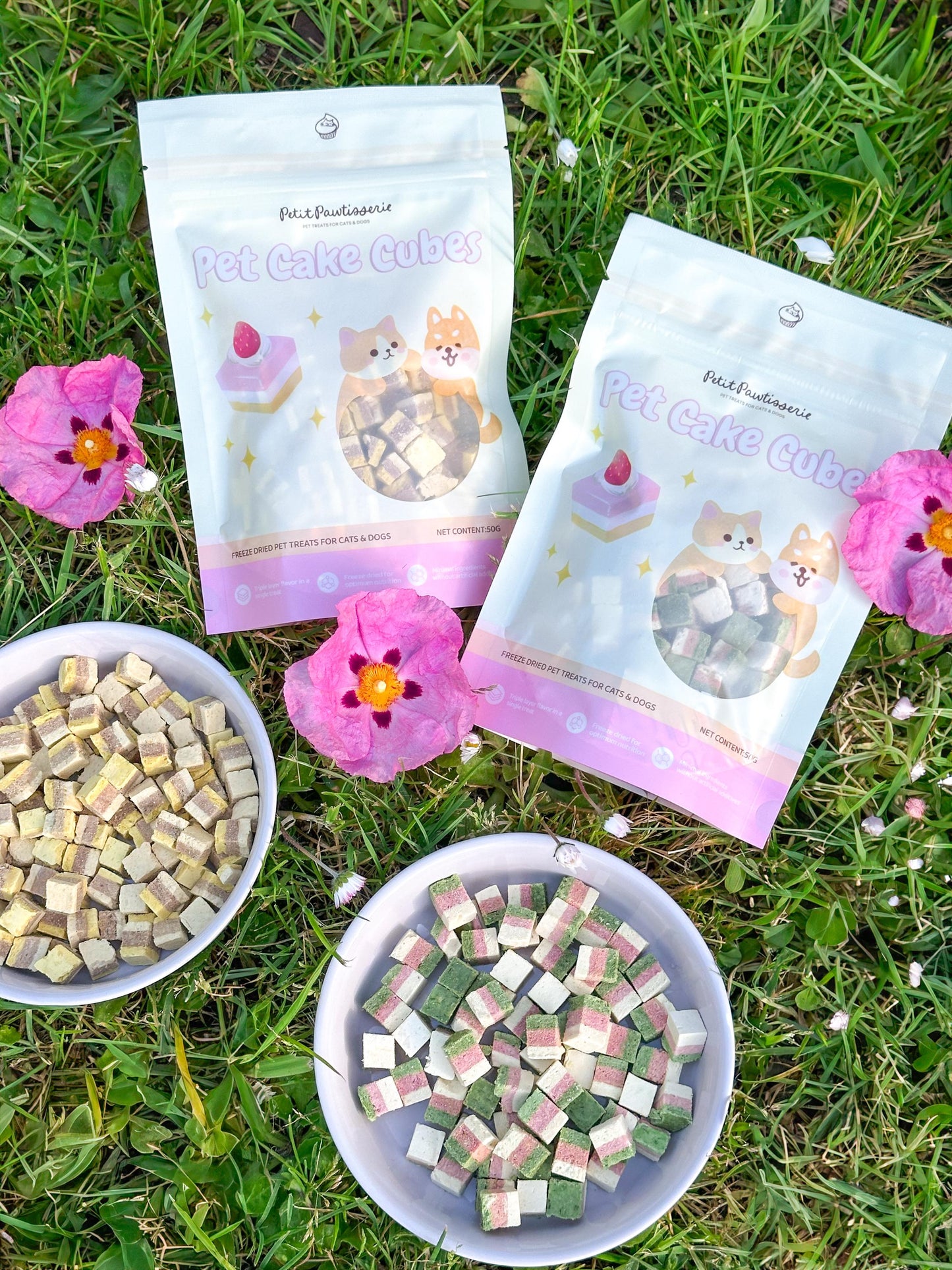Two bags of pet cake cubes, one labeled &#39;Pet Cake Cubes&#39; and the other &#39;Pet Cake Cube.&#39; The bags are on a bed of green grass with pink flowers. The bags are white with pink and yellow accents. The bags feature cartoon images of dogs and cakes. Two bowls of pet cake cubes are in the foreground.  Pawtisserie freeze-dried cake cube pet treats for cats and dogs – soft bite sized snacks with 3 layers of flavor