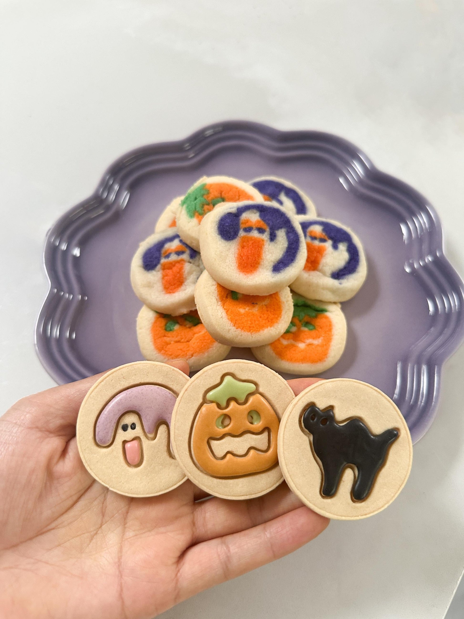 frosted Pilsbury Halloween pet treats on a purple plate, featuring cute ghost, pumpkin, and black cat designs. Festive freeze-dried cookies perfect for pets during the Halloween season.